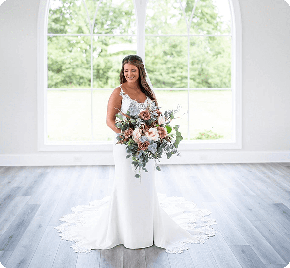 Bride in elegant white gown holding a large floral bouquet indoors.