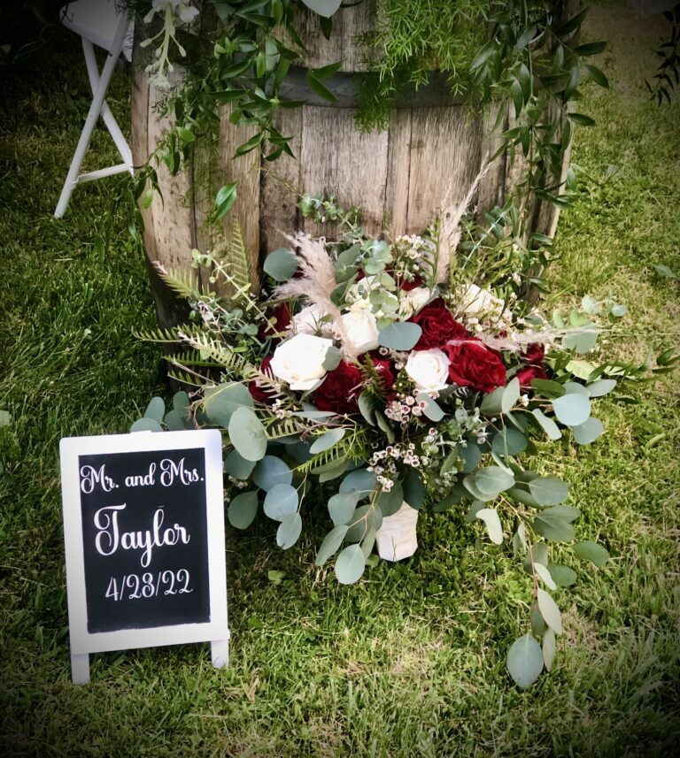 Rustic wedding sign with floral arrangement and greenery beside wooden backdrop.