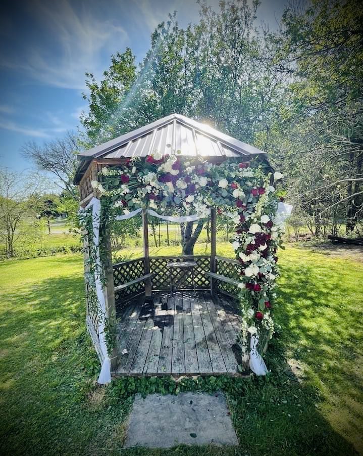 A small wooden gazebo decorated with flowers in a sunlit garden.