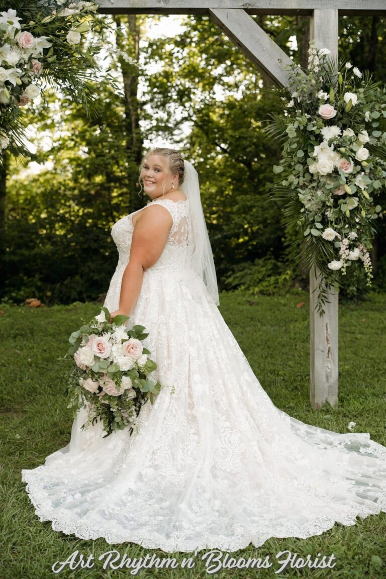 Bride in elegant white wedding gown holding a large bouquet outdoors.