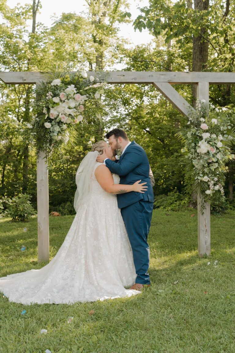 Bride and groom share a kiss under a floral arch outdoors.