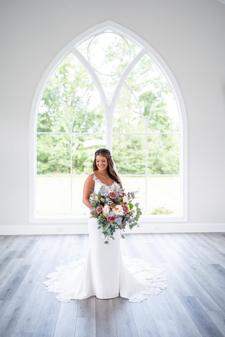 Bride in white dress holding a bouquet in front of a large arched window.