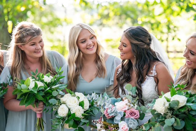 Three women smiling and holding bouquets, dressed in pastel and white dresses outdoors.