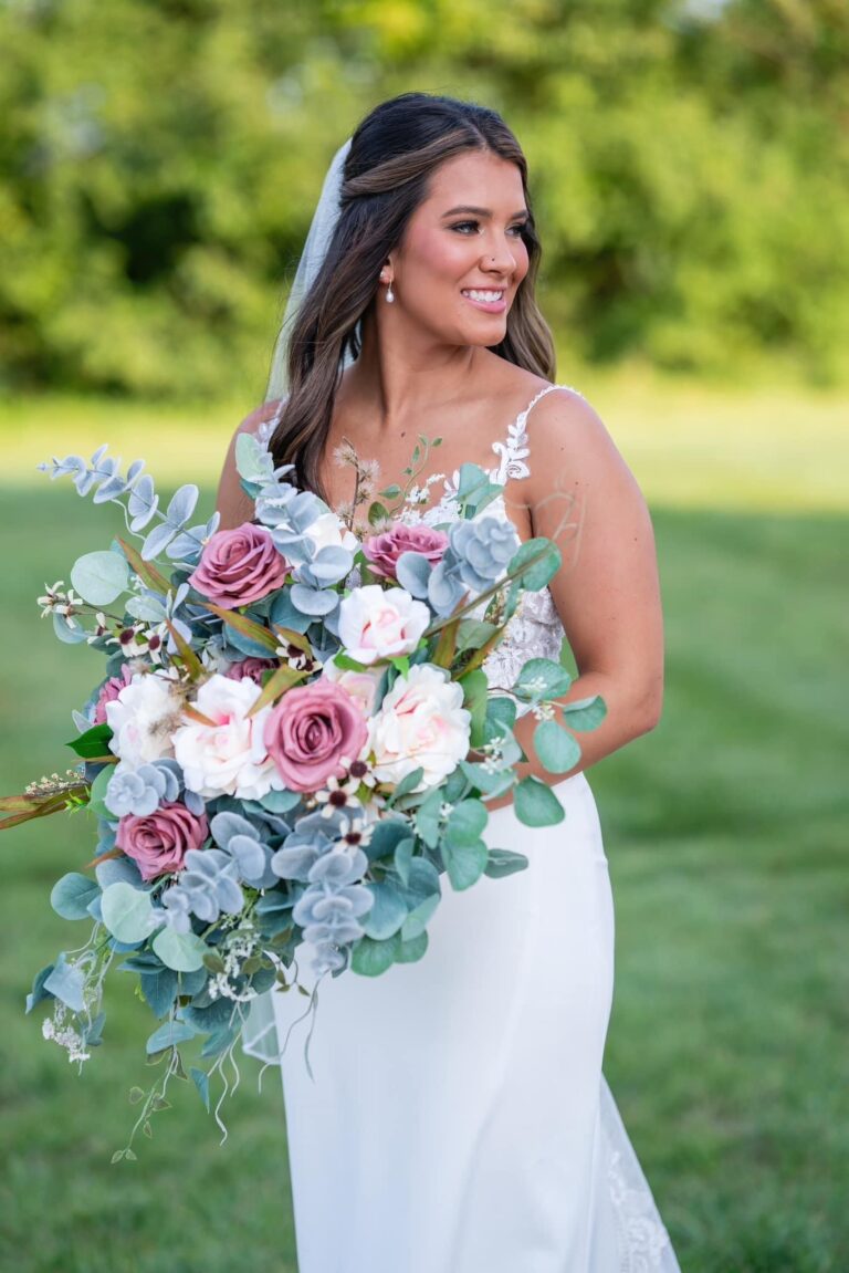 Bride in white dress holding a large floral bouquet outdoors.