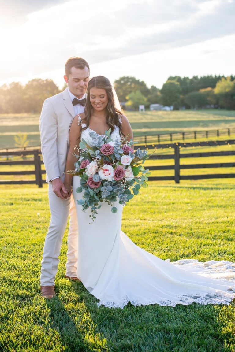 Newlywed couple posing outdoors with a large bridal bouquet in the sunlight.