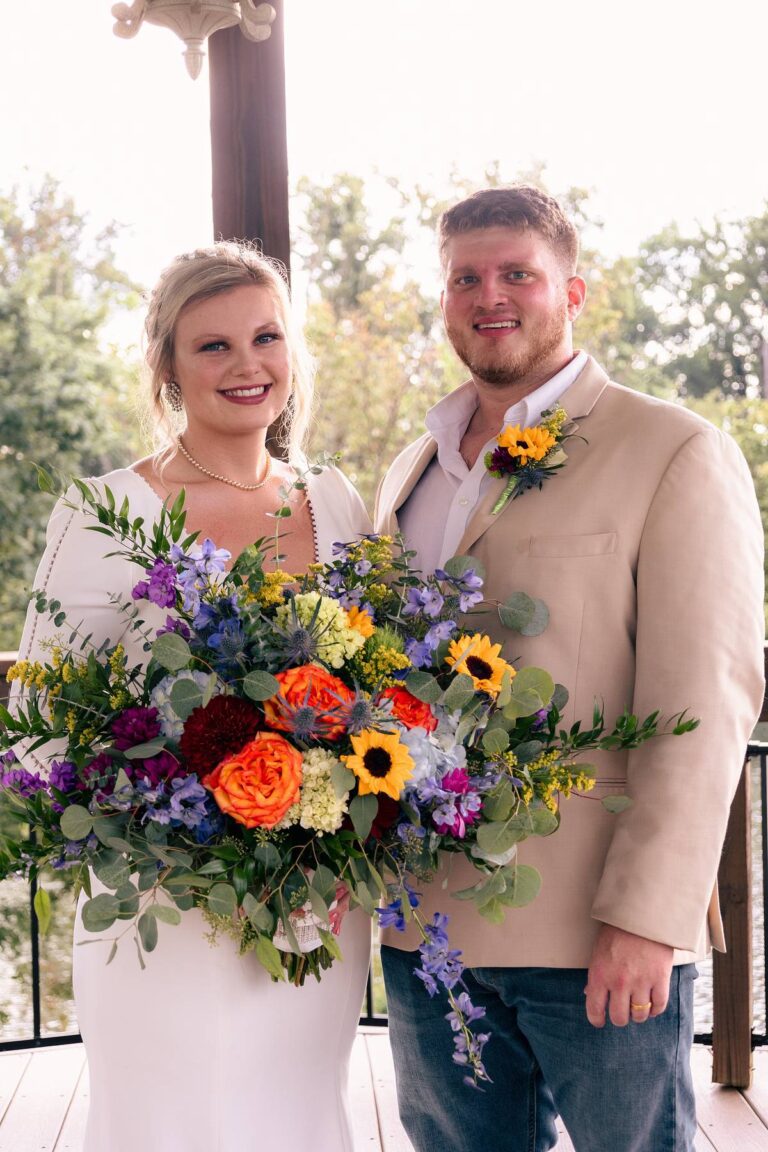 A happy couple in wedding attire holding a colorful bouquet outdoors.
