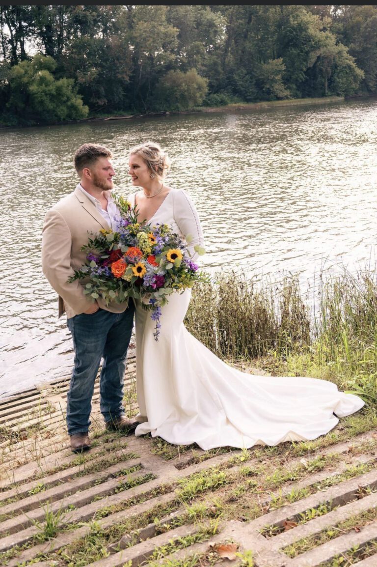 Newlywed couple by the water, holding a vibrant bouquet.