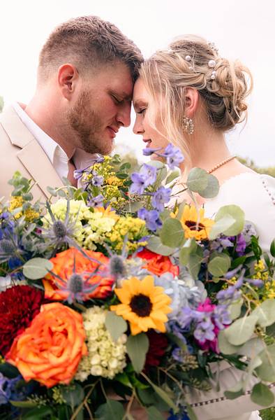 Bride and groom share a tender moment behind a vibrant floral bouquet.